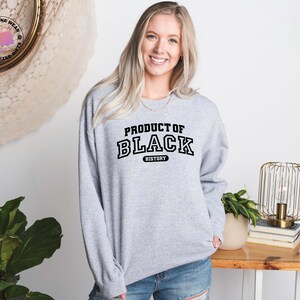 K&ouml;nnte beinhalten: Ein hellgraues Sweatshirt mit dem Text "PRODUCT OF BLACK HISTORY" in schwarzen Buchstaben. Das lang&auml;rmlige Sweatshirt wird von einer Person getragen. Der Hintergrund umfasst einen Holztisch, eine dekorative Lampe und eine Topfpflanze.