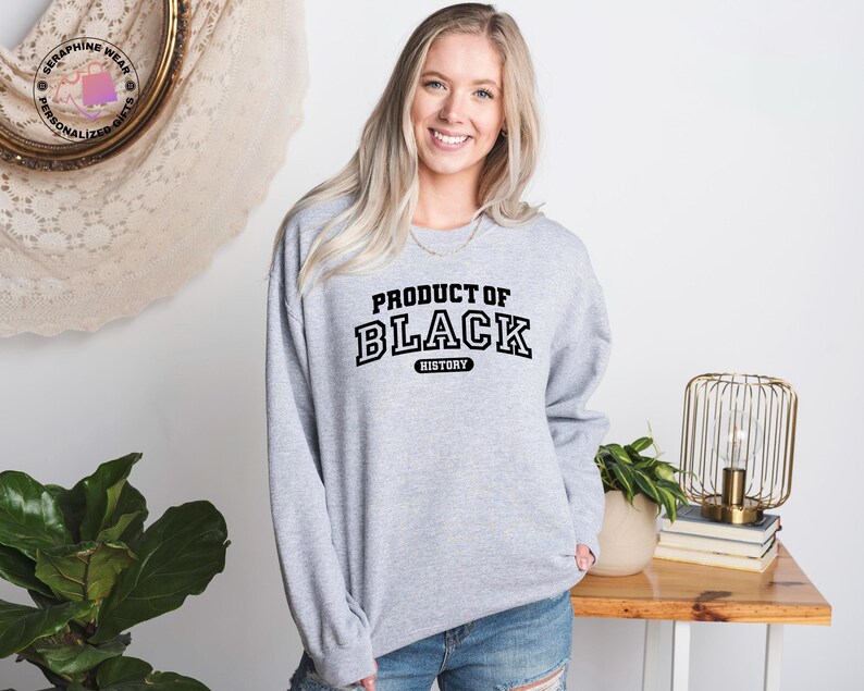 K&ouml;nnte beinhalten: Ein hellgraues Sweatshirt mit dem Text "PRODUCT OF BLACK HISTORY" in schwarzen Buchstaben. Das lang&auml;rmlige Sweatshirt wird von einer Person getragen. Der Hintergrund umfasst einen Holztisch, eine dekorative Lampe und eine Topfpflanze.