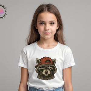May include: A white t-shirt featuring a raccoon wearing a red cap and sunglasses. The text "Feral Kid" is printed on the sunglasses. The shirt is worn by a young person.