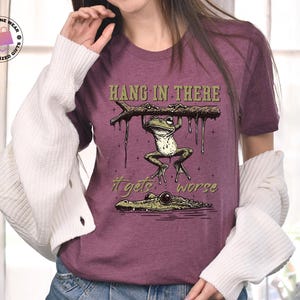 May include: A purple t-shirt with a graphic of a frog hanging from a branch with the text "Hang in there it gets worse".