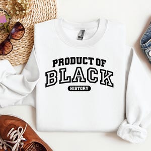 K&ouml;nnte beinhalten: Ein wei&szlig;es Sweatshirt mit dem Text "PRODUCT OF BLACK HISTORY" in fetter, schwarzer Schrift im College-Stil. Das Bild zeigt braune Schuhe, eine Sonnenbrille und Jeans.