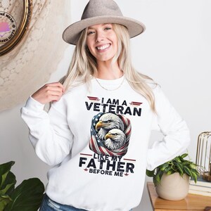 May include: White sweatshirt with the text "I AM A VETERAN LIKE MY FATHER BEFORE ME" and an illustration of two eagles with an American flag design. The person is wearing a grey hat.