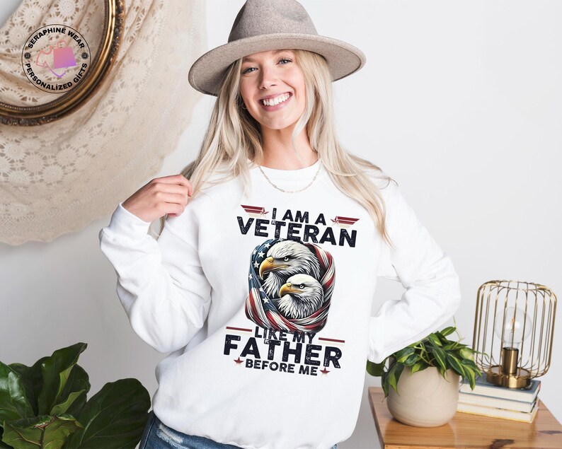 May include: White sweatshirt with the text "I AM A VETERAN LIKE MY FATHER BEFORE ME" and an illustration of two eagles with an American flag design. The person is wearing a grey hat.