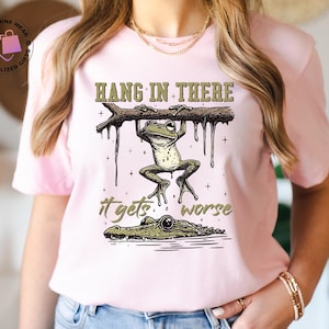 May include: Pink t-shirt with a green frog hanging from a branch with the text "Hang in there it gets worse" and a green alligator below the frog.