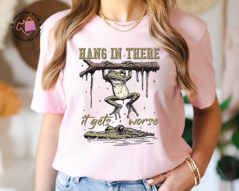 May include: Pink t-shirt with a green frog hanging from a branch with the text "Hang in there it gets worse" and a green alligator below the frog.