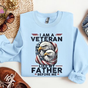 May include: A pale blue sweatshirt with the text "I AM A VETERAN LIKE MY FATHER BEFORE ME." The graphic features two eagles with an American flag design. The sweatshirt is made of a soft material.
