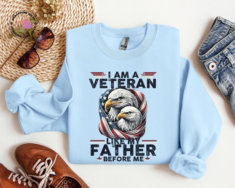 May include: A pale blue sweatshirt with the text "I AM A VETERAN LIKE MY FATHER BEFORE ME." The graphic features two eagles with an American flag design. The sweatshirt is made of a soft material.