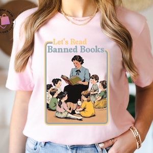 May include: Pale pink t-shirt with a vintage-style illustration of a teacher reading to children, and the text "Let's Read Banned Books". The design is framed in a light blue and yellow border.