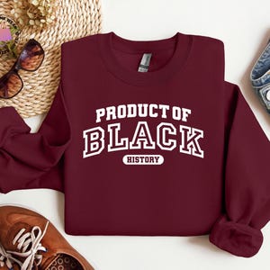 K&ouml;nnte beinhalten: Ein weinrotes Sweatshirt mit dem Text "PRODUCT OF BLACK HISTORY" in wei&szlig;en Blockbuchstaben. Das Sweatshirt wird auf einer wei&szlig;en Oberfl&auml;che mit braunen Schuhen, einer Sonnenbrille und einer Jeans pr&auml;sentiert.
