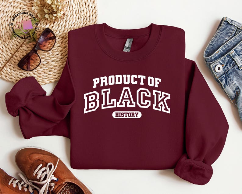 K&ouml;nnte beinhalten: Ein weinrotes Sweatshirt mit dem Text "PRODUCT OF BLACK HISTORY" in wei&szlig;en Blockbuchstaben. Das Sweatshirt wird auf einer wei&szlig;en Oberfl&auml;che mit braunen Schuhen, einer Sonnenbrille und einer Jeans pr&auml;sentiert.