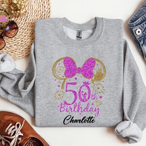 Puede incluir: Una sudadera gris con un lazo rosa brillante y el texto "50th Birthday Charlotte". El dise&ntilde;o incluye detalles dorados y fuegos artificiales. La sudadera es de cuello redondo.