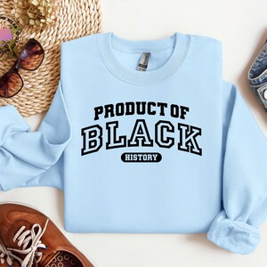 K&ouml;nnte beinhalten: Hellblaues Sweatshirt mit dem Text "PRODUCT OF BLACK HISTORY" in schwarzen Blockbuchstaben. Das Bild zeigt auch braune Schuhe und eine blaue Jeans.