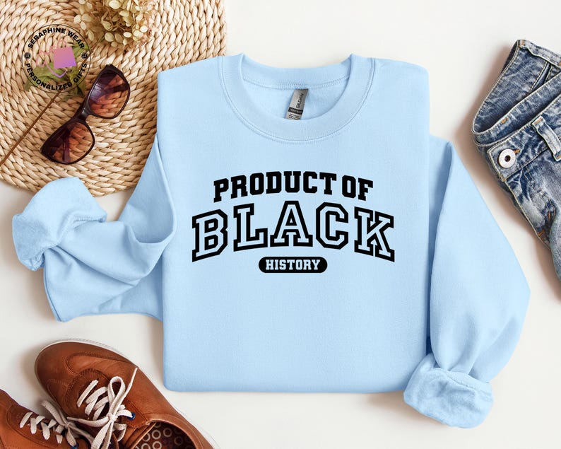 K&ouml;nnte beinhalten: Hellblaues Sweatshirt mit dem Text "PRODUCT OF BLACK HISTORY" in schwarzen Blockbuchstaben. Das Bild zeigt auch braune Schuhe und eine blaue Jeans.