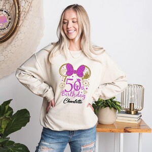 Puede incluir: Sudadera color crema con un lazo morado brillante y dise&ntilde;o de orejas de Minnie Mouse. El gr&aacute;fico incluye el texto "50th Birthday Charlotte" en morado y dorado. Un estilo casual y c&oacute;modo.