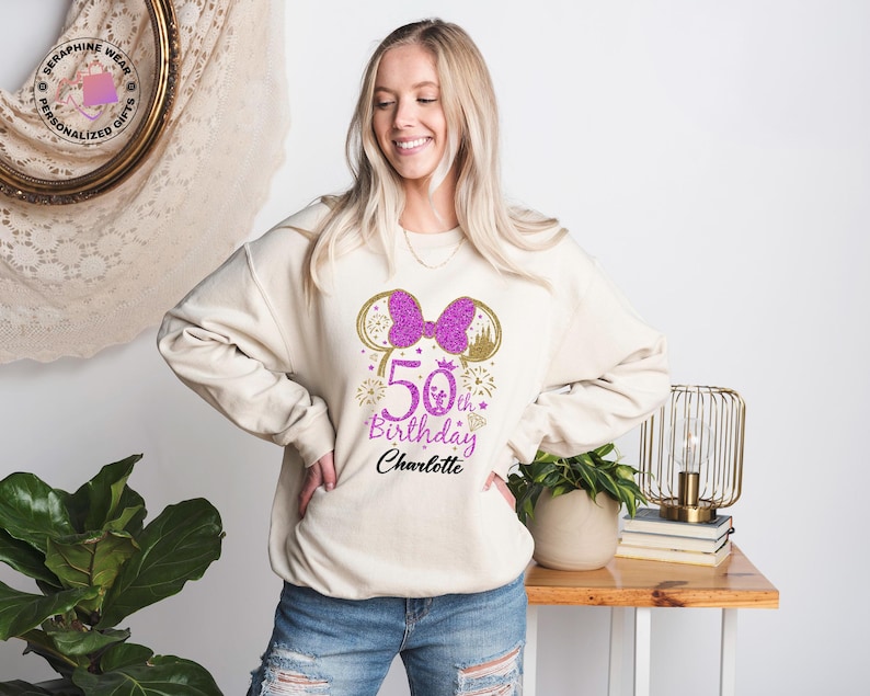 Puede incluir: Sudadera color crema con un lazo morado brillante y dise&ntilde;o de orejas de Minnie Mouse. El gr&aacute;fico incluye el texto "50th Birthday Charlotte" en morado y dorado. Un estilo casual y c&oacute;modo.
