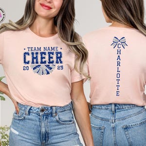 May include: A pale pink t-shirt with the word "CHEER" in blue and silver lettering, and the years "20" and "25". The back of the shirt has a blue bow and the name "CHARLOTTE" in blue lettering.