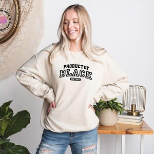 K&ouml;nnte beinhalten: Cremefarbenes Sweatshirt mit dem Text "PRODUCT OF BLACK HISTORY" in schwarzen, fetten Blockbuchstaben. Das Sweatshirt wird von einer Person mit blonden Haaren und Jeans getragen. Der Hintergrund umfasst einen Holztisch, eine Lampe und eine Pflanze.