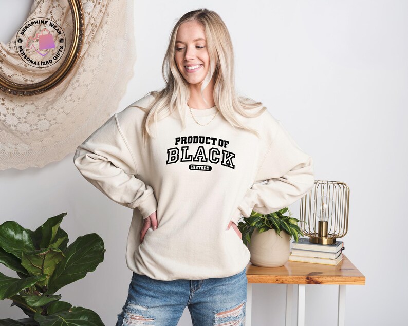 K&ouml;nnte beinhalten: Cremefarbenes Sweatshirt mit dem Text "PRODUCT OF BLACK HISTORY" in schwarzen, fetten Blockbuchstaben. Das Sweatshirt wird von einer Person mit blonden Haaren und Jeans getragen. Der Hintergrund umfasst einen Holztisch, eine Lampe und eine Pflanze.