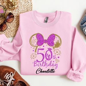 Puede incluir: Una sudadera rosa claro con un lazo morado brillante y un dise&ntilde;o de orejas doradas. El texto "50th Birthday Charlotte" est&aacute; impreso en morado y negro. La sudadera es de cuello redondo.