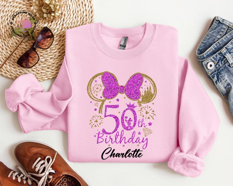Puede incluir: Una sudadera rosa claro con un lazo morado brillante y un dise&ntilde;o de orejas doradas. El texto "50th Birthday Charlotte" est&aacute; impreso en morado y negro. La sudadera es de cuello redondo.