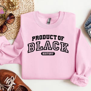 K&ouml;nnte beinhalten: Hellrosa Sweatshirt mit dem Text "PRODUCT OF BLACK HISTORY" in schwarzen Blockbuchstaben. Das Sweatshirt hat einen Rundhalsausschnitt und lange &Auml;rmel. Das Bild zeigt auch eine Sonnenbrille, braune Schuhe und eine Jeans.