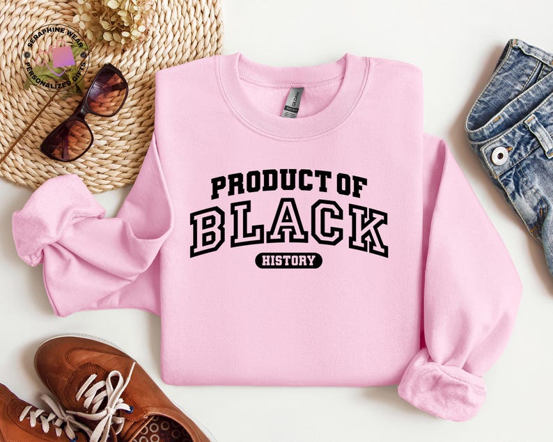 K&ouml;nnte beinhalten: Hellrosa Sweatshirt mit dem Text "PRODUCT OF BLACK HISTORY" in schwarzen Blockbuchstaben. Das Sweatshirt hat einen Rundhalsausschnitt und lange &Auml;rmel. Das Bild zeigt auch eine Sonnenbrille, braune Schuhe und eine Jeans.