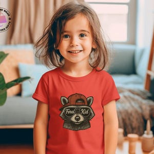 May include: A red t-shirt featuring a raccoon wearing a red cap and sunglasses with the text "Feral Kid". The raccoon graphic is in the center of the shirt. The shirt is being worn by a young child.