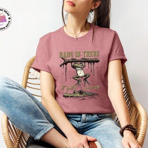 May include: A pink t-shirt with a graphic of a frog hanging on a branch with the text "Hang in there it gets worse".