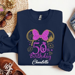 Puede incluir: Sudadera azul marino con un lazo rosa brillante y el texto "50th Birthday Charlotte". El dise&ntilde;o incluye una silueta de castillo dorado, fuegos artificiales y un diamante. La sudadera es de la marca Gildan.