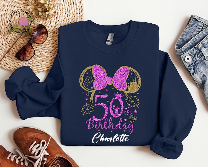 Puede incluir: Sudadera azul marino con un lazo rosa brillante y el texto "50th Birthday Charlotte". El dise&ntilde;o incluye una silueta de castillo dorado, fuegos artificiales y un diamante. La sudadera es de la marca Gildan.