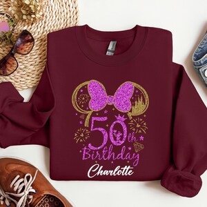 Puede incluir: Sudadera burdeos con un lazo morado brillante y el texto "50th Birthday Charlotte". El dise&ntilde;o incluye detalles dorados, fuegos artificiales y una silueta de castillo. Accesorios rodean la sudadera.