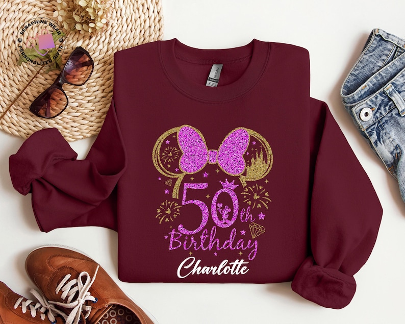 Puede incluir: Sudadera burdeos con un lazo morado brillante y el texto "50th Birthday Charlotte". El dise&ntilde;o incluye detalles dorados, fuegos artificiales y una silueta de castillo. Accesorios rodean la sudadera.