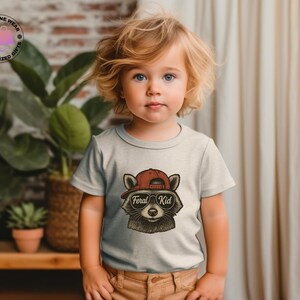 May include: A light gray toddler t-shirt featuring a raccoon wearing a red baseball cap and sunglasses. The text "Feral Kid" is below the raccoon's face. The child is wearing brown pants and is standing in front of a plant.