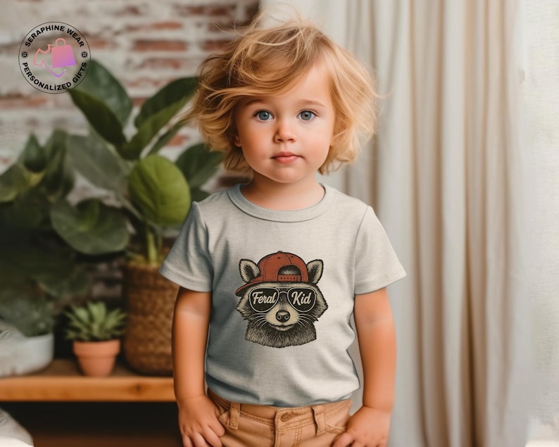 May include: A light gray toddler t-shirt featuring a raccoon wearing a red baseball cap and sunglasses. The text "Feral Kid" is below the raccoon's face. The child is wearing brown pants and is standing in front of a plant.