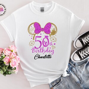Puede incluir: Camiseta blanca con un lazo rosa brillante y un dise&ntilde;o de orejas de rat&oacute;n doradas. La camiseta presenta el texto "50th Birthday Charlotte" en rosa y negro, con fuegos artificiales y un diamante. Zapatillas Converse negras y pantalones cortos vaqueros tambi&eacute;n en la imagen.