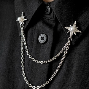May include: Silver starburst brooch with a pearl center and dangling chain detail. The brooch is attached to a black button-down shirt. The starburst design is a classic and elegant accessory.