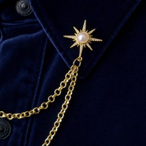 May include: A gold-toned starburst brooch featuring a central white pearl surrounded by small clear crystals. Two gold-toned metal chains hang from the bottom of the starburst, attached to a dark blue velvet fabric with visible buttons.