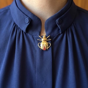 Könnte beinhalten: Eine goldfarbene Käferbrosche mit roten Akzenten ist an einem dunkelblauen Hemd mit Kragen befestigt. Die Brosche zeigt ein Käferdesign. Das Hemd hat einen geknöpften Kragen und einen weichen, fließenden Stoff.