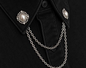 May include: A black collared shirt featuring two silver-toned metal collar pins. Each pin is adorned with a central oval faux pearl surrounded by small clear crystals. A double-layered silver chain connects the two pins across the front.