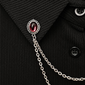 Op de afbeelding: Een zilverkleurige broche met een ovale, donkerrode cabochonsteen in een sierlijke filigraan omlijsting. Twee fijne zilverkleurige metalen kettingen hangen aan de basis van de broche, wat zorgt voor een gelaagd, gedrapeerd effect op de stof.