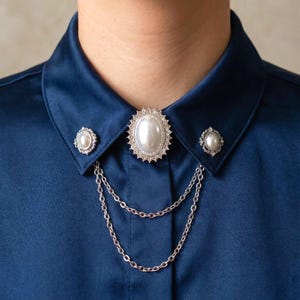 May include: A navy blue satin shirt with a decorative brooch set. The set includes a large oval pearl-like centerpiece surrounded by a silver-colored, crystal-studded frame, two smaller matching brooches, and a double chain.