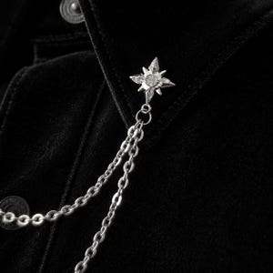 Może przedstawiać: Srebrzysta przypinka do klapy w kształcie czteroramiennej gwiazdy z małymi osadzonymi kryształkami. Przypinka jest połączona z dwoma delikatnymi srebrzystymi metalowymi łańcuszkami, które zwisają w dół na czarnej aksamitnej powierzchni.