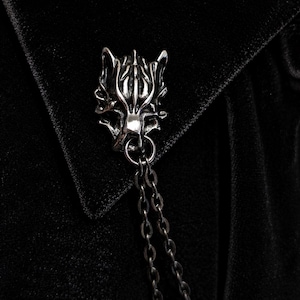 May include: A silver-toned metal brooch shaped like a stylized wolf head, featuring intricate carved details and a polished finish. Two thin, dark metal chains are attached to a small ring at the base of the wolf head, hanging downward in a parallel line.