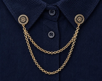 May include: A gold-toned metal collar pin featuring two circular discs with a floral embossed design, connected by two draped cable chains. The accessory is attached to the collar points of a dark navy blue ribbed button-down shirt.