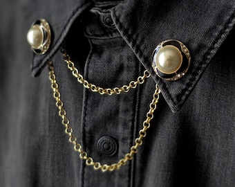 May include: A black denim shirt featuring a collar adorned with two circular pins. Each pin has a central cream-colored faux pearl surrounded by a black enamel border and a gold-tone rim. Two gold-tone metal chains connect the pins, draping across the front of the shirt placket.