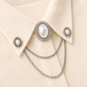 May include: A cream-coloured corduroy shirt collar adorned with three silver-toned brooches featuring oval faux pearls surrounded by small clear crystals. The brooches are connected by two draped silver-toned metal chains hanging below the collar.