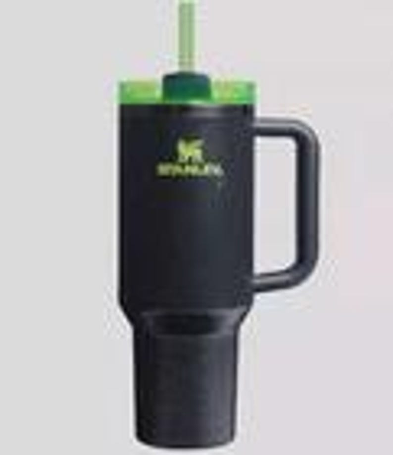 Op de afbeelding: Zwarte Stanley-beker met een groene deksel en rietje. De beker heeft een handvat en het Stanley-logo in groen. De beker is ontworpen om drankjes koud of warm te houden. De beker is ongeveer 23 cm hoog.