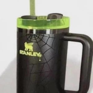 Op de afbeelding: Zwarte Stanley reismok met een groene deksel en handvat. De mok heeft een spinnenwebontwerp en het Stanley-logo in geel. Een groen rietje is in het deksel gestoken.