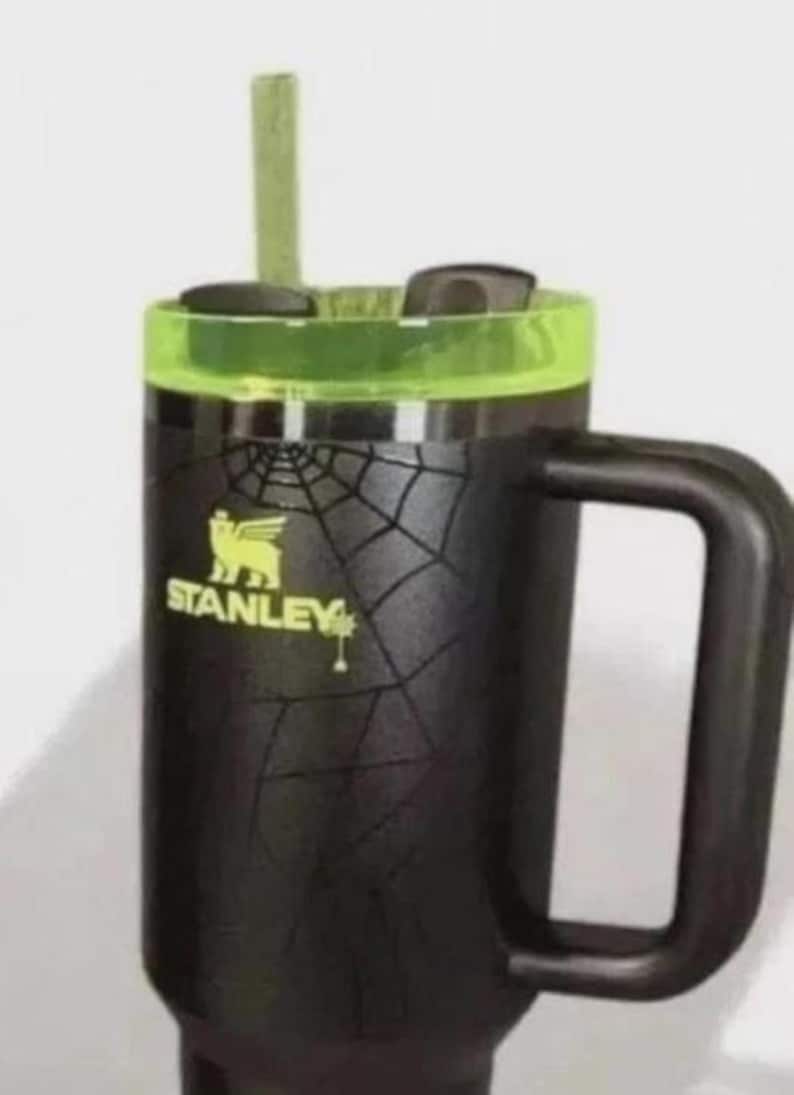 Op de afbeelding: Zwarte Stanley reismok met een groene deksel en handvat. De mok heeft een spinnenwebontwerp en het Stanley-logo in geel. Een groen rietje is in het deksel gestoken.