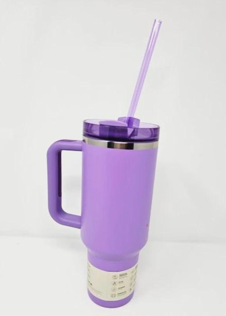 May include: A large, purple insulated tumbler with a handle and a clear straw. The tumbler has a lid and a silver band near the top. The bottom of the tumbler has a white label with icons.